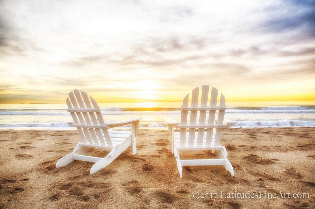Two white Adirondack chairs on a beach facing a stunning yellow-blue sunset.