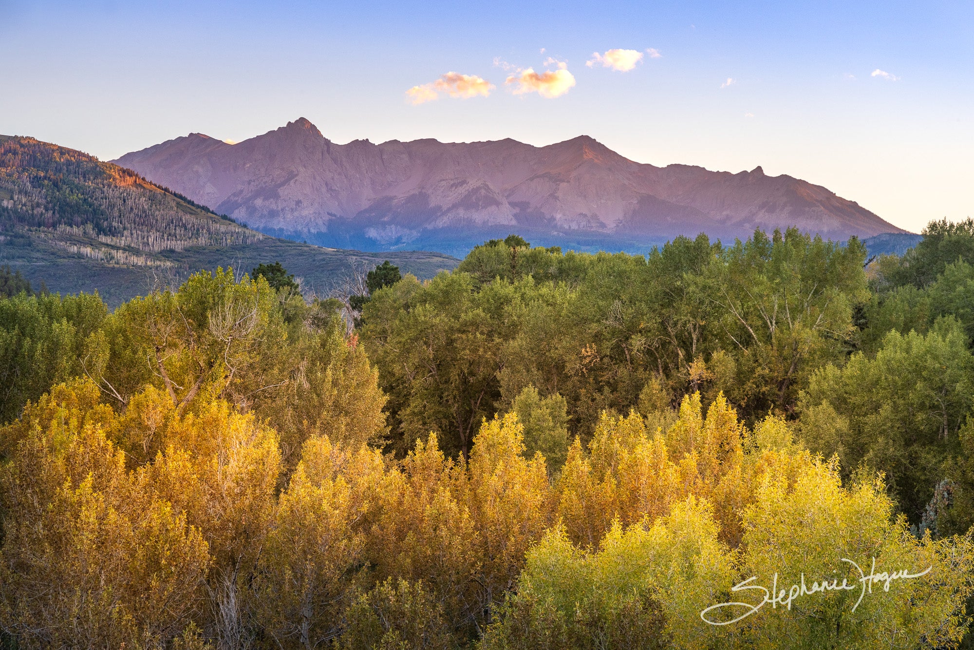 Fine art nature photography of of a setting sun providing a blanket of soft light over fall-colored trees and a dramatic mountain silhouette.